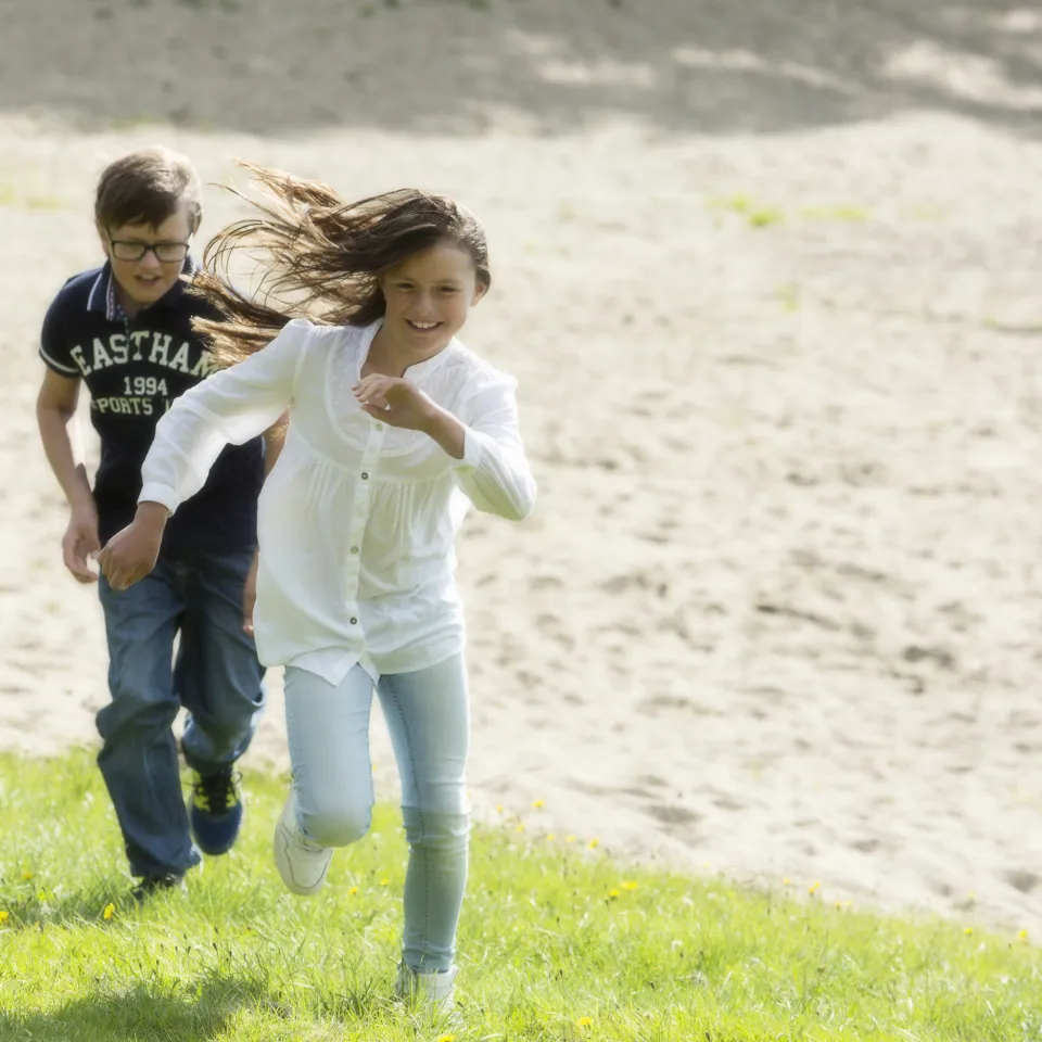 Två barn springer uppför en grässluttning i solsken, med sandig yta i bakgrunden och vind i håret.
