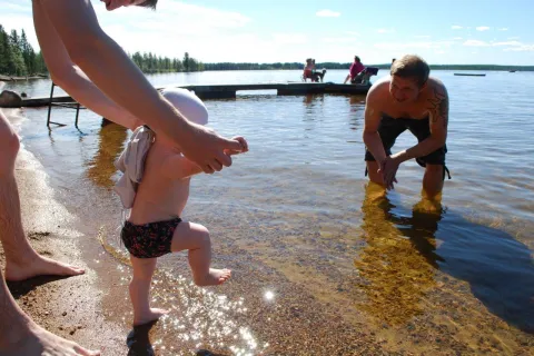 Ett litet barn leds av en vuxen ner i grunt sjövatten vid en strand, med brygga och badgäster i bakgrunden.