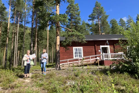 Röd timrad trästuga med vit ramp bland tallar i soligt väder, med två personer på en stig i förgrunden.