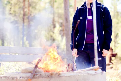 En person i mörk jacka står vid en brinnande eldstad utomhus, med rök och träd i bakgrunden.