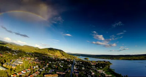 Flygvy över Malå vid Malån, med skogsklädda kullar, blå himmel med moln och en regnbåge.