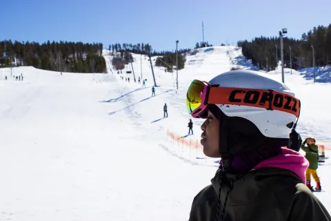 Närbild av skidåkare med vit hjälm och skidglasögon i förgrunden, med snöiga skidbackar och åkare i bakgrunden.