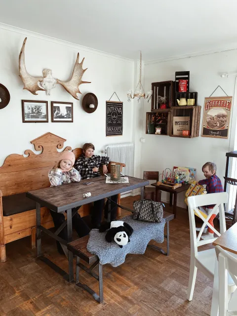 Barn som har fikapaus under shoppingtur vid rustik matplats med träbord, bänk och stolar, väggmonterade horn, tavlor och hyllor med lådor och pynt.