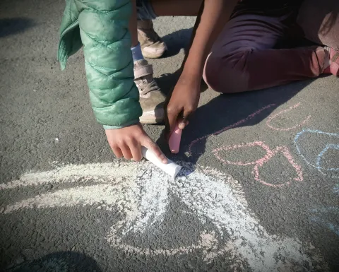Två barn sitter på asfalt och ritar med kritor; händer, skor och färgglada kritlinjer syns i solljus.